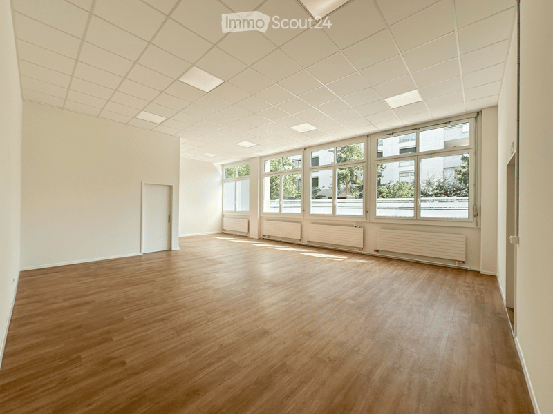 Empty room with wooden flooring, white walls, large windows, radiators, and a white door.