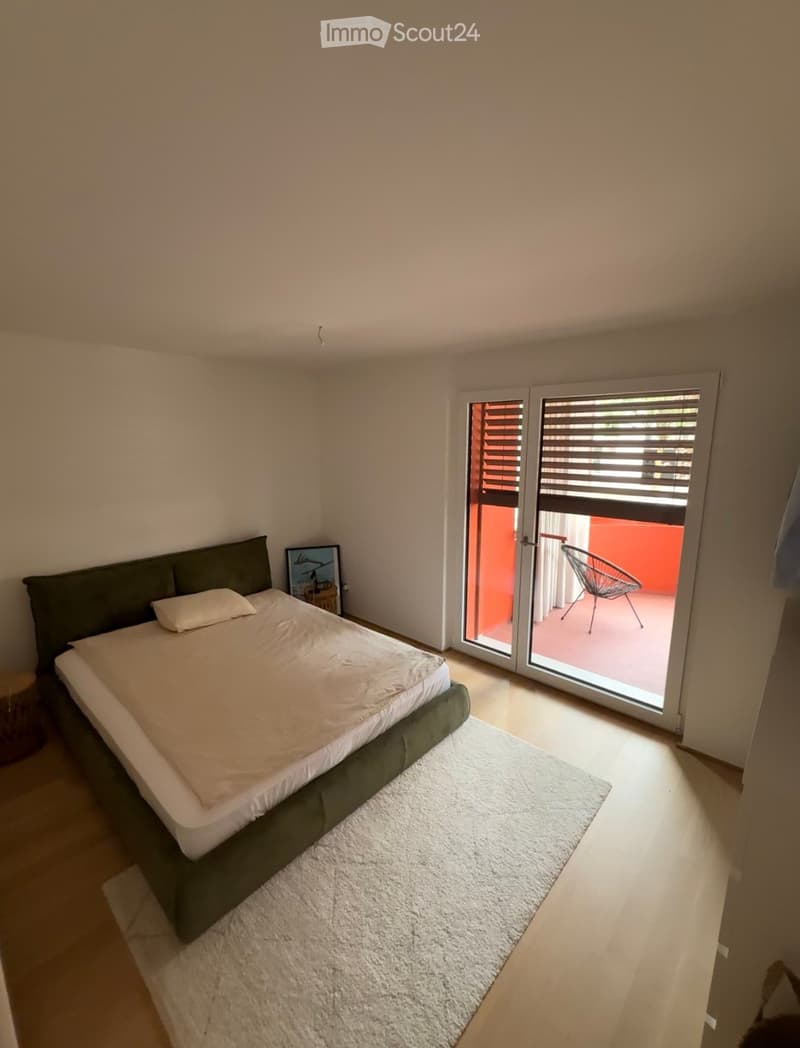 Modern bedroom with a large bed, white bedding, a white rug, and a sliding glass door leading to a balcony with a chair.