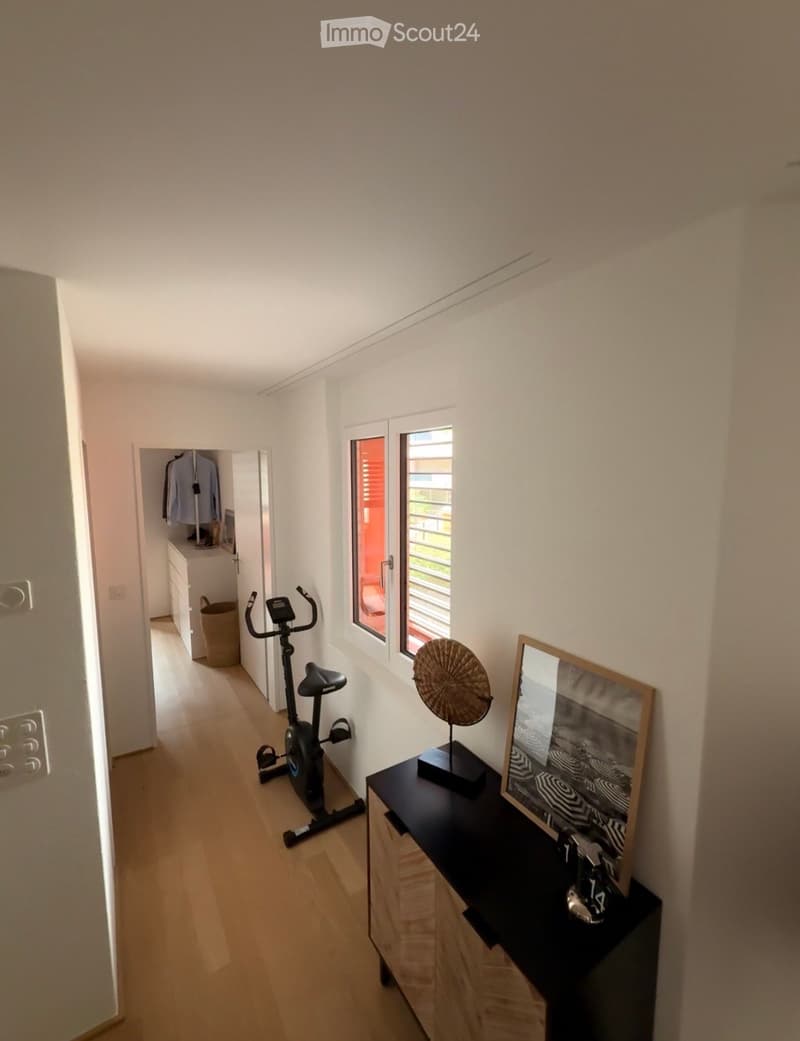 Hallway with wooden flooring, exercise bike, small table with a framed picture and decorative object, and a closet with clothes visible inside.