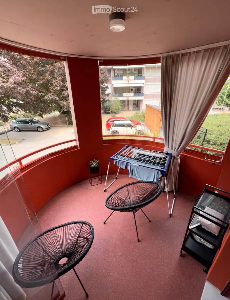 Balcony with red walls, pink carpet, two black wire chairs, small table with folded clothes, potted plant, and curtains.