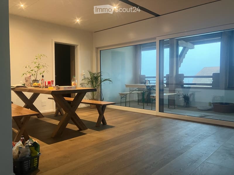 Bright living space with wooden flooring, large glass sliding doors leading to a patio, a wooden table with benches, potted plants, and various items on the table.