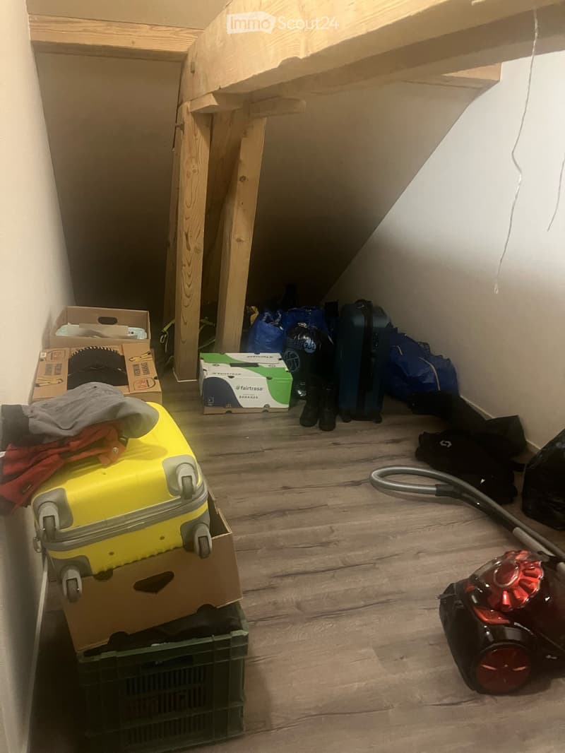 A cluttered room with wooden flooring, containing a yellow suitcase, vacuum cleaner, cardboard boxes, and various bags.