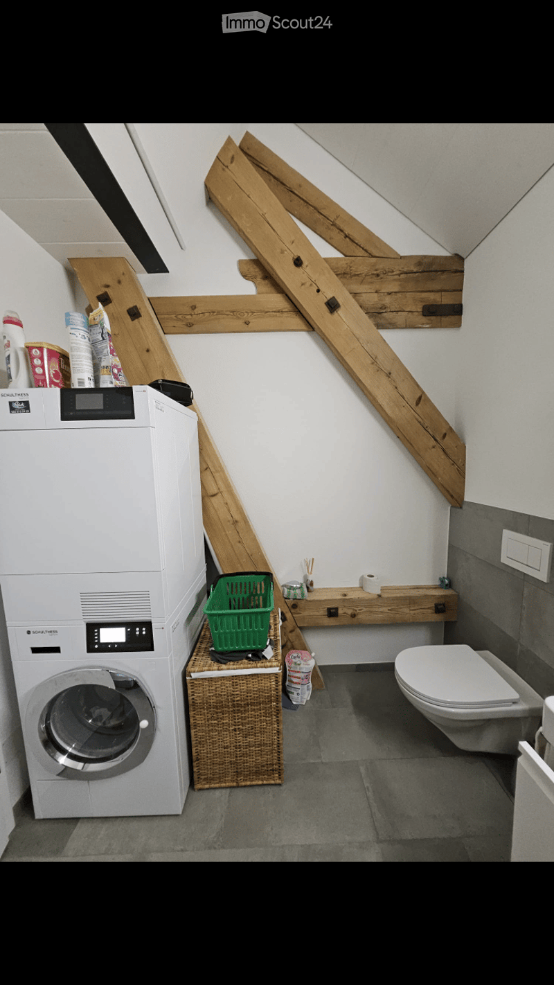 Bathroom with a washing machine, toilet, wooden shelf, and a small woven basket on the floor.