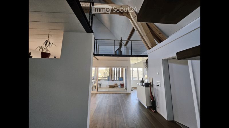 Open, modern hallway with wooden flooring, white walls, and a glass door leading to a living area. Features a metal railing and a small plant on a shelf.