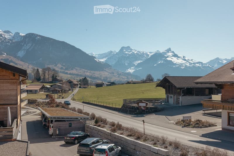 A scenic view of a village with traditional wooden houses, a parking area, and snow-capped mountains in the background.