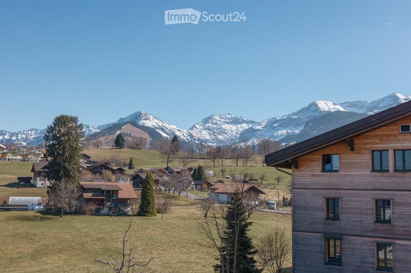 A wooden house with large windows overlooking a scenic valley and snow-capped mountains in the background.