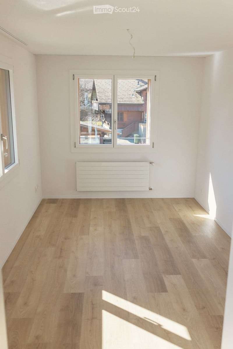 Empty room with light wooden flooring, white walls, and a large window offering a view of a neighboring house and roof. A white radiator is mounted on the wall below the window.
