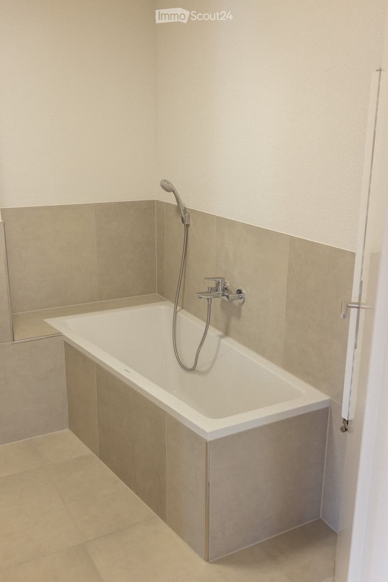 Modern bathroom with a white bathtub, beige tiled walls, and a showerhead attached to the wall.