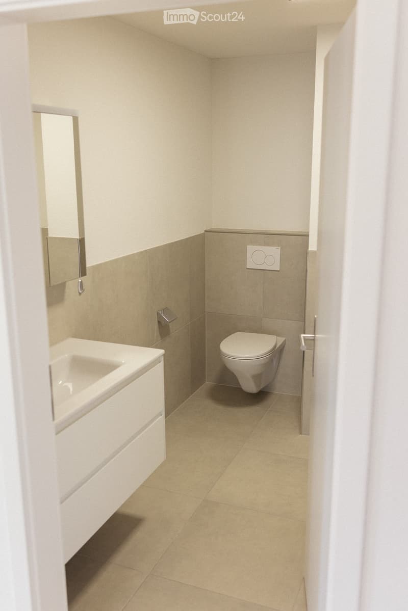 Modern bathroom with a white sink, toilet, and mirror, featuring beige tiled walls and floor.