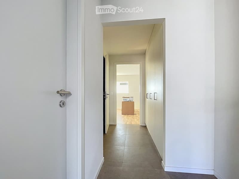 A clean, modern hallway with tiled flooring, white walls, and a door on the left leading to another room. The hallway has a simple design with a visible cabinet and a small table in the background.