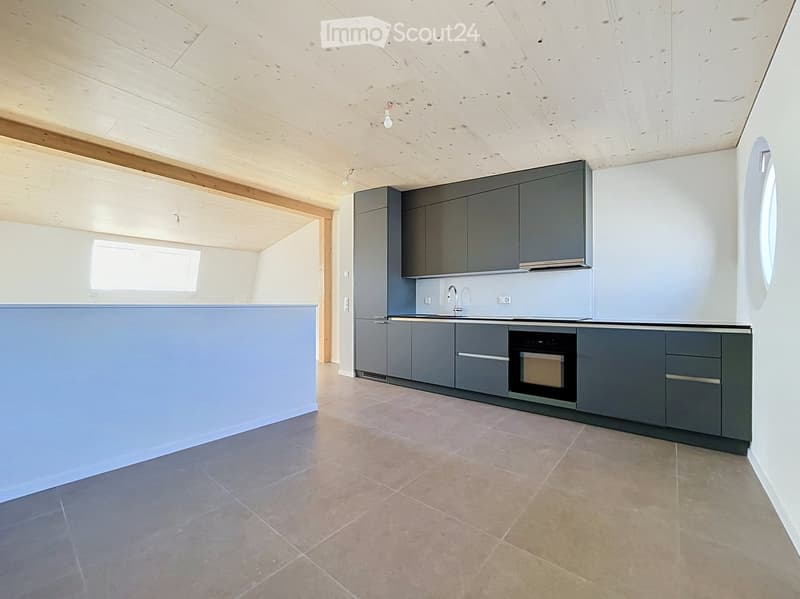 Modern kitchen with grey cabinets, a central island, and a built-in oven. The space features light wooden beams on the ceiling and large windows allowing natural light.