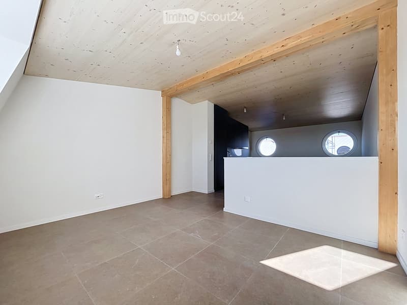 An empty attic space with exposed wooden beams, tiled floor, and white walls. Features a small white counter with two round windows above it.