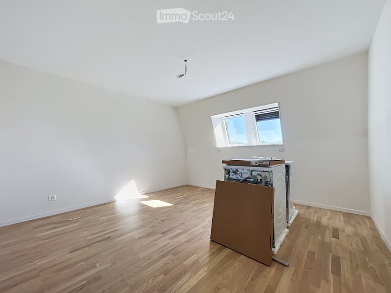 Empty room with wooden flooring, white walls, and a window with blinds. A washing machine is partially visible behind a cardboard cover.