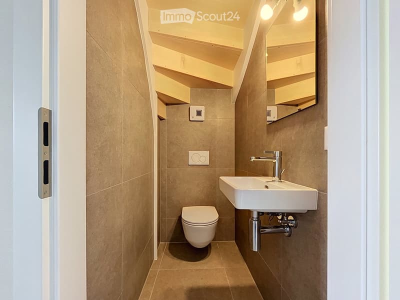 Modern, compact bathroom with beige tiled walls and floor, featuring a toilet, a white sink with a chrome faucet, and a mirrored cabinet above the sink.