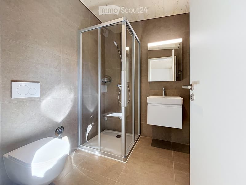 Modern bathroom with a glass-enclosed shower, toilet, sink, and mirror. Features beige tiles and a sleek design.