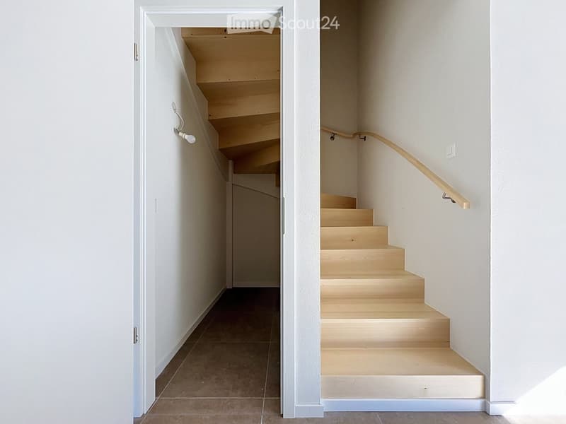 Modern hallway with a wooden staircase featuring a light-colored wooden railing and steps, white walls, and a tiled floor.