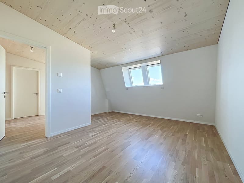 Empty room with wooden flooring, white walls, exposed wooden ceiling, and a single window letting in natural light.
