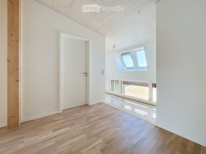 Empty room with wooden flooring, white walls, and large windows with blinds. The room features a white door and exposed wooden beams on the ceiling.