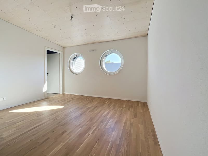 Empty room with wooden flooring, white walls, and two round windows offering a view. The ceiling is made of light wood panels.