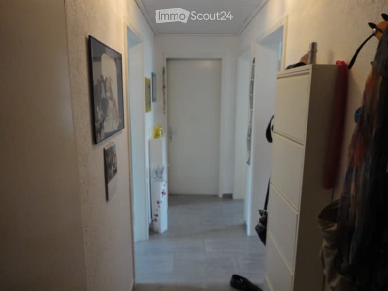 A narrow hallway with tiled flooring, white walls, and several doors. There are framed pictures on the walls and a small cabinet with items on top.