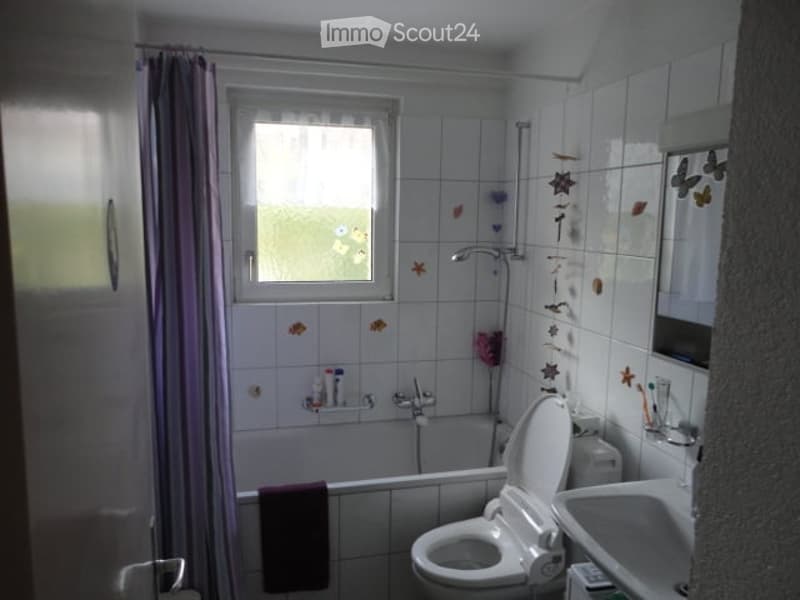 A small bathroom with white tiled walls and floor, featuring a bathtub, toilet, sink, and shower. A window with a curtain allows natural light to enter, and decorative tiles with butterfly and star motifs adorn the walls.