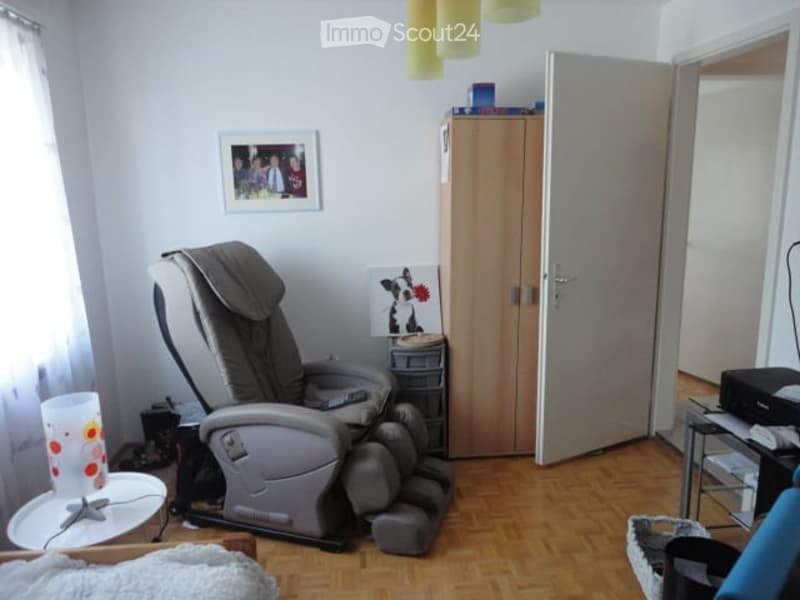 A cozy room with a wooden floor, a large recliner chair, a wardrobe, a small table with a lamp, and a picture frame on the wall.