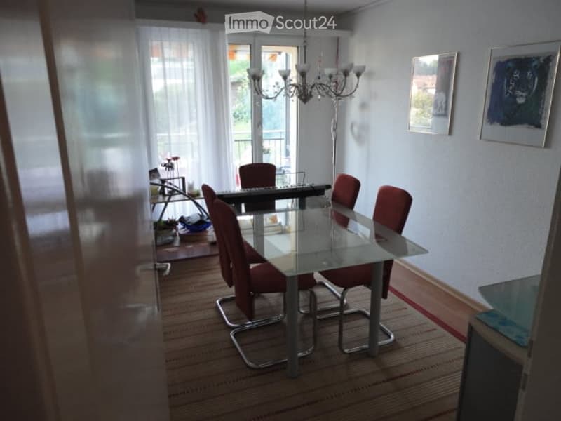 Dining room with a glass table, red chairs, chandelier, and large windows with a view to a balcony.