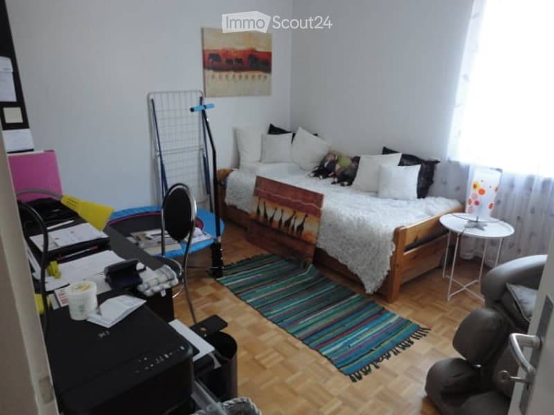 Cozy, well-furnished room with a sofa, bed, chair, small table, lamp, and artwork. Wooden flooring and colorful rug.