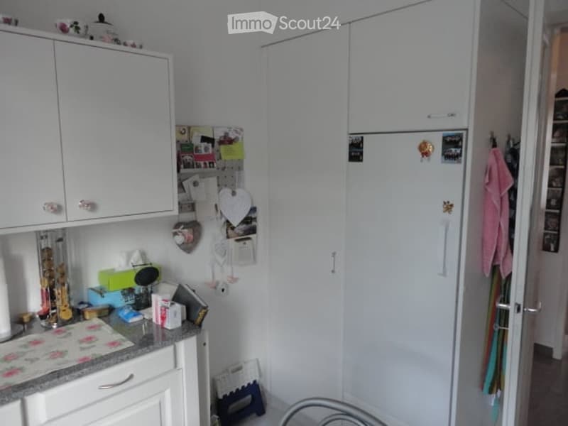 A compact kitchen with white cabinets, a refrigerator, and a countertop filled with various kitchen items, including utensils, a calendar, and a towel hanging on the door.