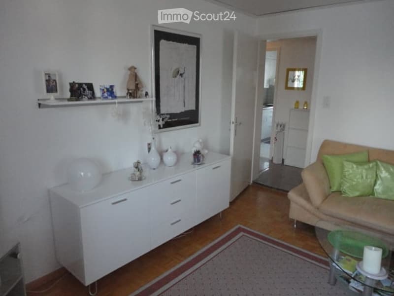 A modern living room with a white cabinet, a beige sofa with green cushions, a glass coffee table, and decorative vases on the cabinet. The room features wooden flooring and a patterned rug.