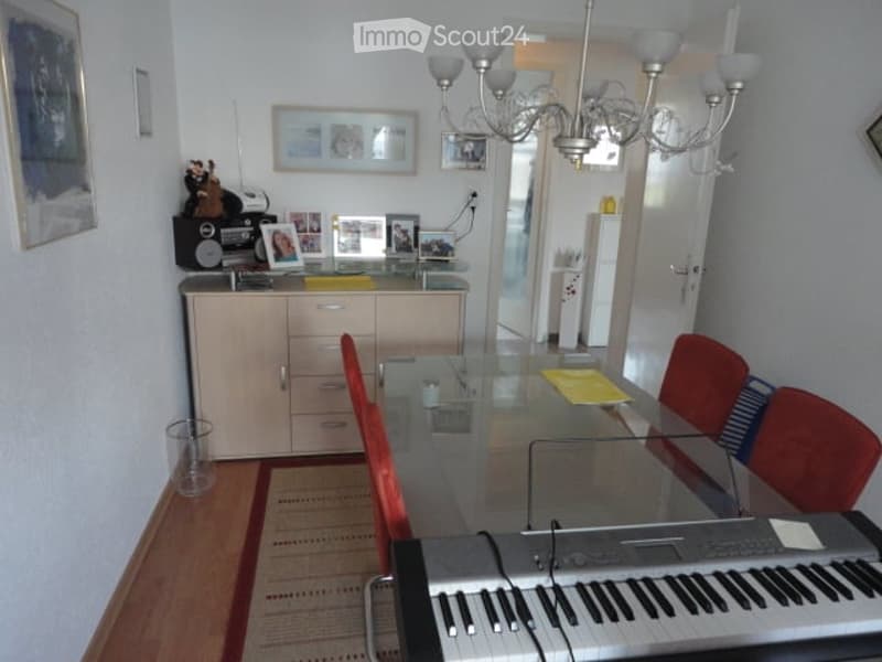 Dining room with a glass table, red chairs, a cabinet, and a keyboard. The room features a chandelier and several picture frames on the wall.
