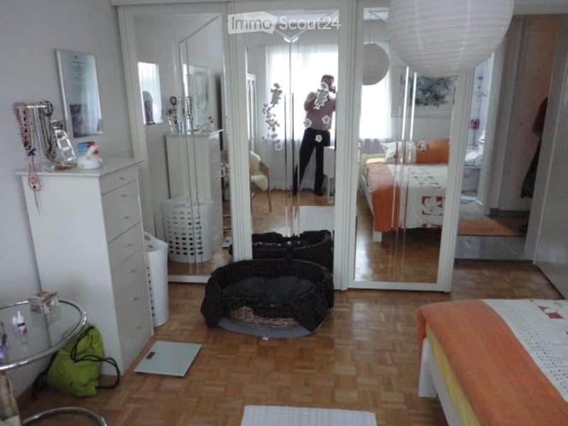 A well-lit bedroom with wooden flooring, a large mirrored wardrobe, a bed with orange and white bedding, a small table, and a chair. The room also features a pet bed and a small round glass table.