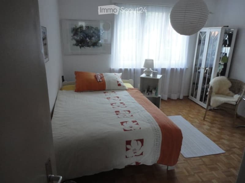Cozy bedroom with a neatly made bed, nightstand with lamp, a small chair, and a mirror. The room features wooden flooring and large windows with white curtains.