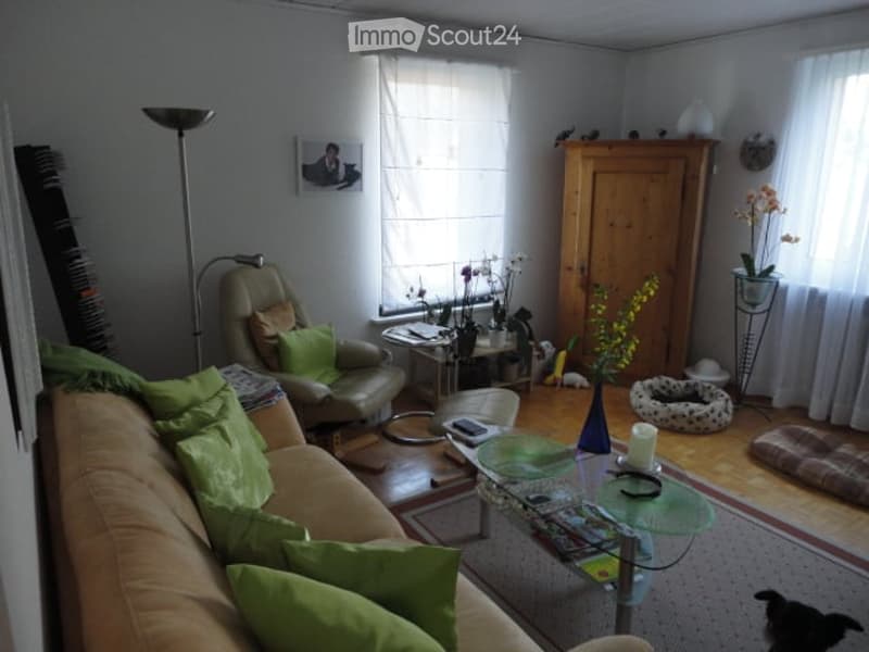 Living room with a beige sofa, green cushions, a glass coffee table, armchair, floor lamp, small table with a vase of flowers, and a picture frame on the wall.