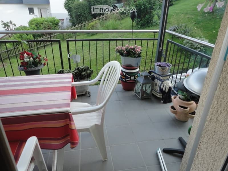 Balcony with tiled floor, metal railing, and several potted plants, including flowers and succulents. There is a white plastic chair and a small table with a striped tablecloth.