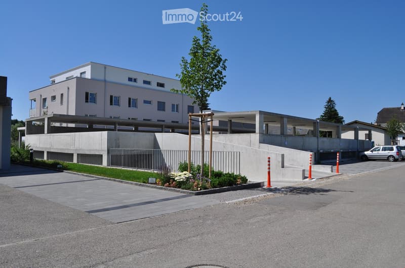 Modern two-story building with a raised ground floor, concrete facade, and a parking area with a covered section.