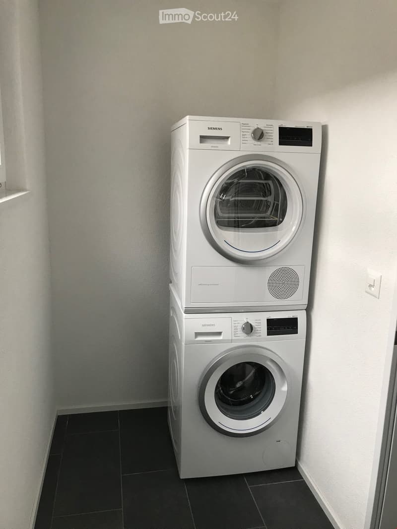 A compact bathroom with a stacked Siemens washing machine and dryer unit against a white wall, featuring a dark tiled floor.