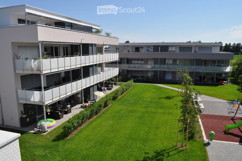 Modern multi-story residential building with balconies, surrounded by a well-maintained lawn, playground, and outdoor seating area.