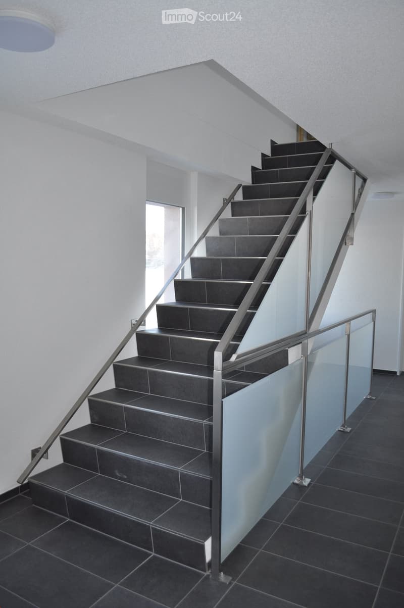 Modern staircase with dark tiled steps and a sleek metal railing featuring a glass panel, set against white walls and a recessed ceiling light.