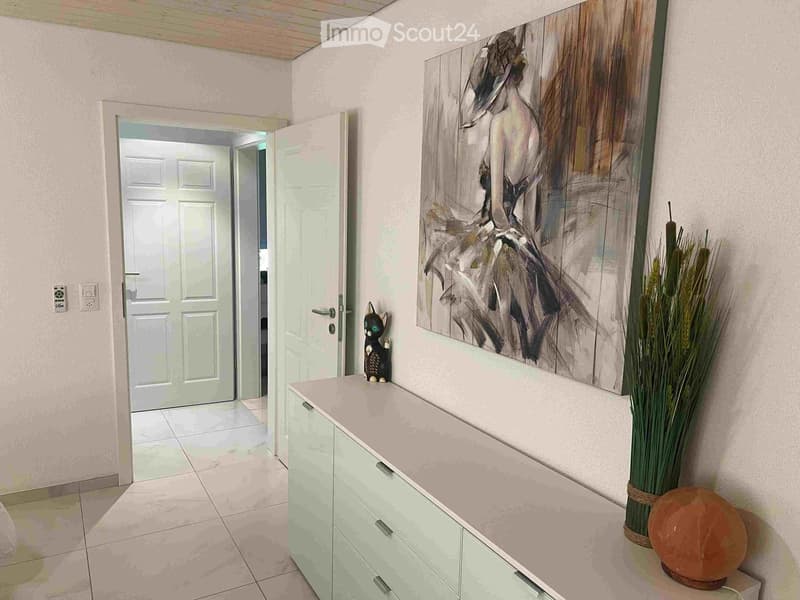 A modern hallway with light-colored walls, tiled flooring, and a large abstract painting on one wall. A small cabinet with drawers is positioned against the wall, topped with a decorative cat figurine and a tall vase with dried plants. An open door leads