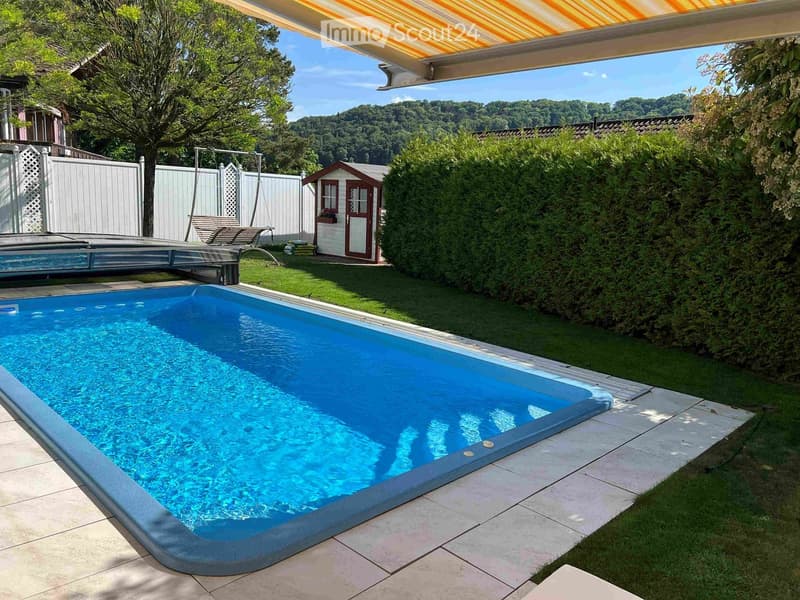 A private garden with a swimming pool, surrounded by green grass, trees, and a small shed. The pool is rectangular with a tiled deck and a covered patio area nearby.