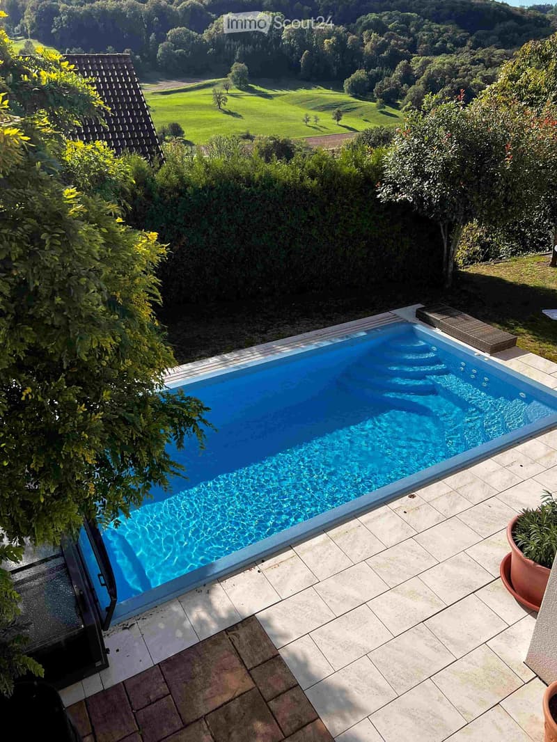 A modern swimming pool with clear blue water, surrounded by a tiled patio and lush greenery, with a scenic view of a green field and trees in the background.