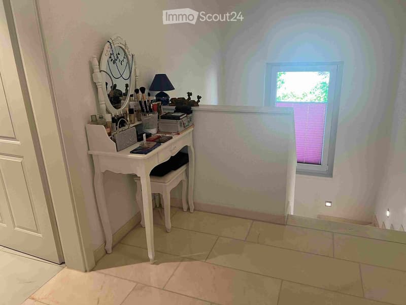 A small, well-lit room with a vanity table, mirror, stool, lamp, and various cosmetics and brushes on the table. The room has tiled flooring and a window with blinds.