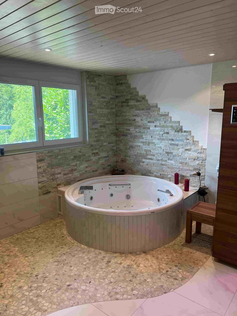 Modern bathroom with a large, round bathtub, stone tiled walls, and a small wooden stool beside it.