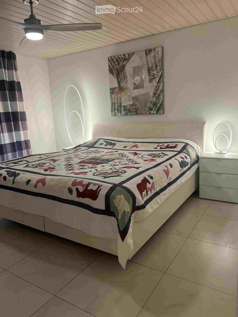 A cozy bedroom with a large bed covered in a colorful quilt, a nightstand with a lamp, a mirror on the wall, and a ceiling fan with light fixture.