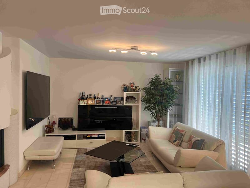 Modern living room with a white sofa, coffee table, ottoman, TV stand, and decorative shelf. Large windows with sheer curtains and a potted plant add to the bright ambiance.
