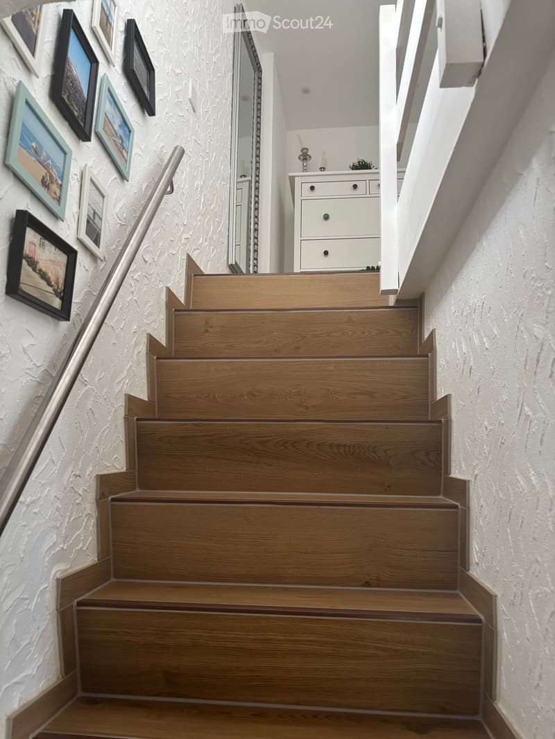 Wooden staircase with a metal handrail, white textured walls, and framed pictures on one side. A small white cabinet with drawers and a mirror is visible at the top of the stairs.
