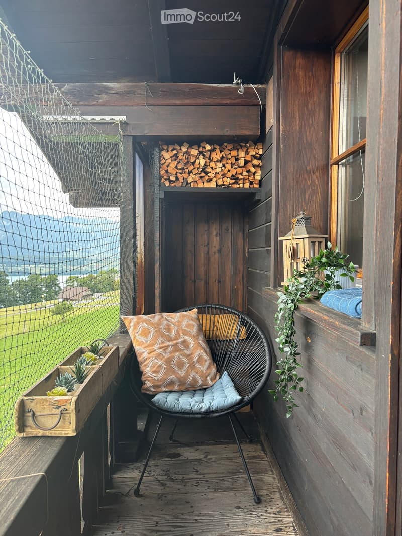 Wooden balcony with a view, featuring a black wire chair with a cushion, a wooden crate with plants, and a wooden shelf with firewood.