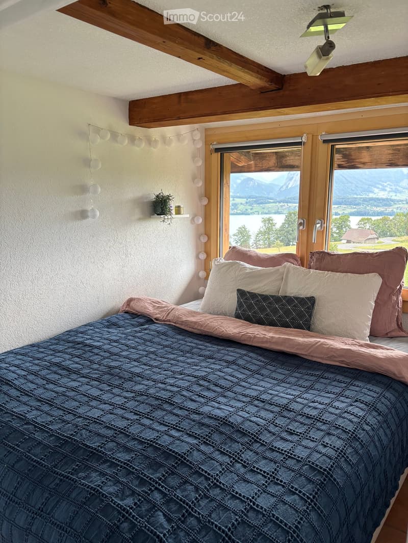 Cozy bedroom with a large window offering a scenic mountain and lake view, featuring a neatly made bed with pillows and a blanket, a small plant on a shelf, and string lights on the wall.