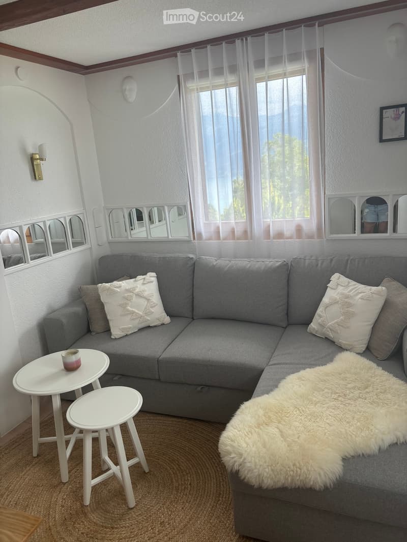 A modern living room with a grey sectional sofa, white cushions, a fur throw, two white stools, a small white table with a candle, and sheer white curtains on the windows.
